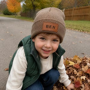 May include: A light brown knit beanie featuring a rectangular faux leather patch on the front cuff. The patch is tan with the name BEN embossed in black capital letters. The beanie has a ribbed texture and a folded cuff design.