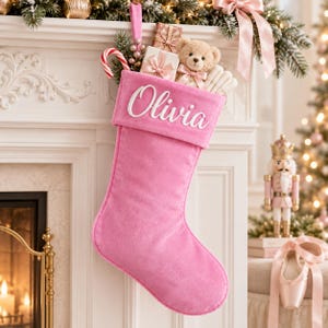 Op de afbeelding: Een zachte roze kerstkous van fluweel met de naam Olivia in wit schrift op de boord geborduurd. De kous is gevuld met een kleine teddybeer, een zuurstok en ingepakte cadeautjes en hangt aan een witte schoorsteenmantel.