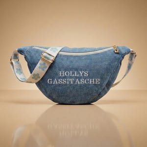 Könnte beinhalten: Eine Jeans-Bauchtasche mit einem weißen Riemen mit blauen Blumenmustern. Die Tasche hat eine geschwungene Form, einen goldenen Reißverschluss und den gestickten Text "HOLLYS GASSITASCHE" auf der Vorderseite.