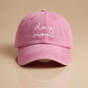 Könnte beinhalten: Rosa Baseballkappe mit dem gestickten Text "dog mom" in weißer Schrift. Die Kappe hat einen gebogenen Schirm und ein sechsteiliges Design. Der Stoff hat ein leicht verblasstes Aussehen.