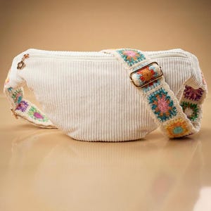 Könnte beinhalten: Eine weiße Cord-Bauchtasche mit einem bunten gehäkelten Riemen. Der Riemen weist quadratische Blumenmuster in Rosa-, Blau-, Orange- und Grüntönen auf. Die Tasche hat einen goldenen Reißverschluss und Beschläge.