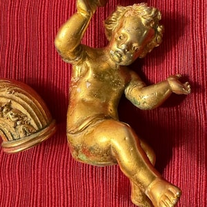 Peut inclure: Une statue de chérubin doré tenant un tambourin, avec un aspect vieilli et détaillé. Le chérubin est posé dans une position assise dynamique. Le fond est un tissu rouge texturé. Art décoratif.