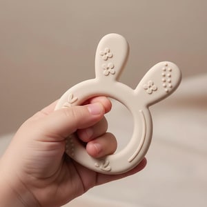 May include: A beige baby teether in the shape of a bunny with floral and dot details. The teether is held in a hand. The teether is made of a soft, flexible material. The bunny ears have floral details, and the other ear has dot details.