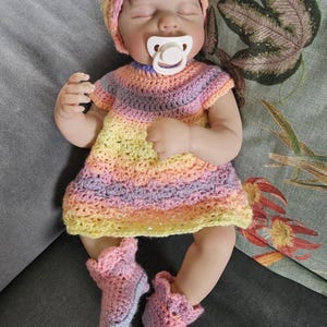 Puede incluir: Una mu&ntilde;eca beb&eacute; con un conjunto de ganchillo. La mu&ntilde;eca tiene un chupete en la boca y viste un vestido a rayas, un gorro y botines en tonos amarillos, rosas y morados. La mu&ntilde;eca est&aacute; tumbada sobre una superficie gris.