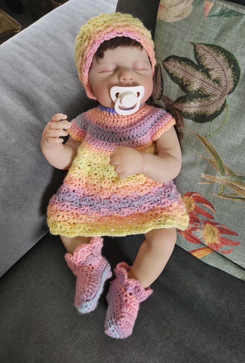 Puede incluir: Una mu&ntilde;eca beb&eacute; con un conjunto de ganchillo. La mu&ntilde;eca tiene un chupete en la boca y viste un vestido a rayas, un gorro y botines en tonos amarillos, rosas y morados. La mu&ntilde;eca est&aacute; tumbada sobre una superficie gris.