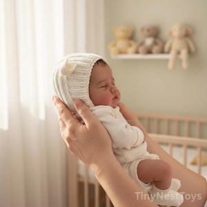 Puede incluir: Un beb&eacute; reci&eacute;n nacido durmiendo, con un gorro blanco de punto con un detalle floral, sostenido en las manos. El beb&eacute; viste un body blanco. El fondo incluye una cuna y una estanter&iacute;a con peluches.