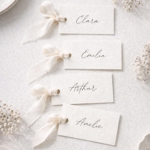 Puede incluir: Cuatro tarjetas de mesa marfil con los nombres Clara, Emilia, Arthur y Amelie escritos en elegante escritura. Cada tarjeta está adornada con un lazo de cinta color crema. Las tarjetas están dispuestas sobre una superficie blanca, con detalles florales y un plato.