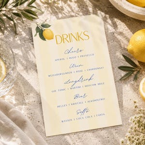 Könnte beinhalten: Eine Getränkekarte mit dem Wort "DRINKS" in goldener Schrift, mit einer Zitronenillustration. Die Karte listet Getränkekategorien wie "Cheers", "Wein", "Longdrink", "Bier" und "Softs" auf. Ein Glas Wasser mit einer Zitronenscheibe und eine ganze Zitrone sind ebenfalls zu sehen.