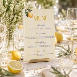 Könnte beinhalten: Eine Menükarte mit einer Zitronenillustration und dem Wort "MENU" in goldener Schrift. Die Karte listet Aperitif, Vorpeise, Hauptgang und Dessert auf. Die Karte wird von einem goldfarbenen Metallständer gehalten, mit Zitronen und Blumen im Hintergrund.