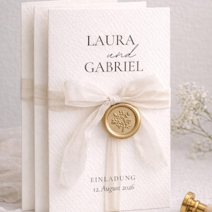 Könnte beinhalten: Elfenbeinfarbene Hochzeitseinladungskarten mit den Namen "LAURA und GABRIEL" in Schwarz gedruckt. Die Karten sind mit einem durchsichtigen Band gebunden und mit einem goldenen Wachssiegel versehen. Die Einladung enthält auch das Datum "12. August 2026".