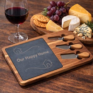 Peut inclure: Planche à fromage en bois avec insert en ardoise, trois couteaux à fromage et le texte "Our Happy Home". La planche est entourée de fromage, de crackers et de raisins. Un verre de vin rouge est en arrière-plan.