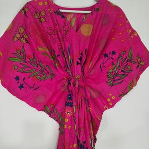 May include: A bright pink kaftan featuring a V-neckline and a central tie detail. The fabric is decorated with a botanical print in shades of green, yellow, and dark blue, including leaves, berries, and sun-like shapes. It hangs on a white hanger.