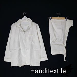 May include: A white pyjama set consisting of a long-sleeved button-down shirt with a collar and a chest pocket, paired with matching white trousers featuring an elastic waistband and a drawstring. The set is displayed on two separate hangers.