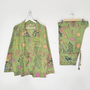 May include: A sage green pyjama set featuring a long-sleeved button-down shirt with a collar and a chest pocket, paired with matching trousers. The fabric is decorated with a colourful botanical print of various flowers, leaves, and sun shapes.