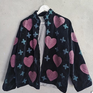 May include: Black jacket featuring embroidered pink hearts and teal crosses. The jacket has a high collar and a striped lining. It is hanging on a white hanger against a grey background.