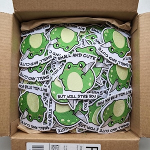 May include: A collection of die-cut stickers featuring a green frog holding a silver knife. The text on each sticker reads "SMALL AND CUTE BUT WILL STAB YOU" in black capital letters. The stickers are packed inside a brown cardboard shipping box.