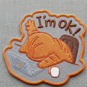 May include: Embroidered patch featuring an orange cat lounging in a chair in front of a laptop. The patch has the words "I'm ok!" in brown. A white mug is next to the laptop. The patch is outlined in orange.