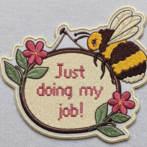 May include: Embroidered patch featuring a cartoon bumblebee perched above a sign. The sign reads "Just doing my job!" and is surrounded by pink flowers and green leaves. The bee has yellow and brown stripes.