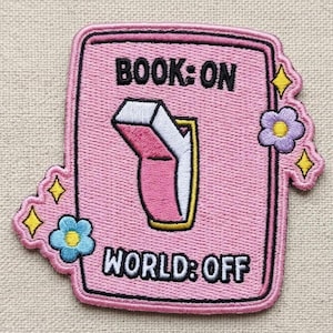 May include: Pink embroidered patch with the text "BOOK: ON" above a switch graphic and "WORLD: OFF" below. The switch is in the "ON" position, with a book graphic. The patch is decorated with yellow stars and blue and purple flowers.