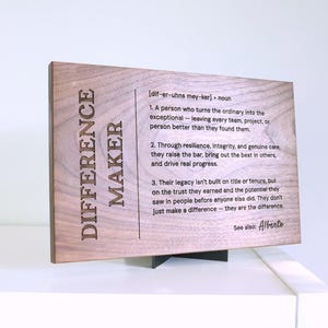 May include: A rectangular wooden plaque with a dark brown grain, featuring the laser-engraved text "DIFFERENCE MAKER" and a dictionary-style definition. The plaque is supported by a small black stand, displaying the name "Alberto" at the bottom.