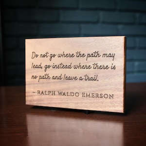 May include: A rectangular wooden sign with a smooth, light brown finish. The text is engraved in a dark, legible font, reading: Do not go where the path may lead, go instead where there is no path and leave a trail. - RALPH WALDO EMERSON.