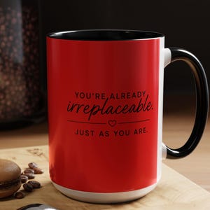 May include: A red and white ceramic mug with a black handle and interior. The mug features the text "YOU'RE ALREADY irreplaceable, JUST AS YOU ARE." A spoon, coffee beans, and a macaron are on a wooden surface.