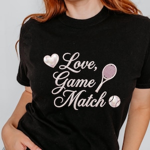 Puede incluir: Camiseta negra con el texto "Love, Game, Match" en fuente cursiva, acompañada de un corazón, una raqueta de tenis y una pelota. El diseño es en blanco y rosa.