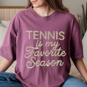 Puede incluir: Camiseta de color malva con la frase "TENNIS is my Favorite Season" en letras perladas. La camiseta de manga corta tiene un corte holgado. El texto es el elemento principal del diseño.