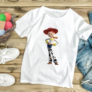 May include: White t-shirt featuring a full-color graphic of the character Jessie from Toy Story. Jessie is wearing a red hat, yellow top, blue jeans with black and white cow spots, and brown boots. The shirt is on a wooden surface.
