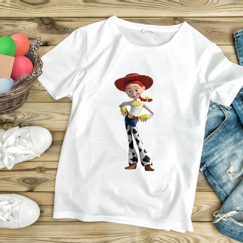 May include: White t-shirt featuring a full-color graphic of the character Jessie from Toy Story. Jessie is wearing a red hat, yellow top, blue jeans with black and white cow spots, and brown boots. The shirt is on a wooden surface.