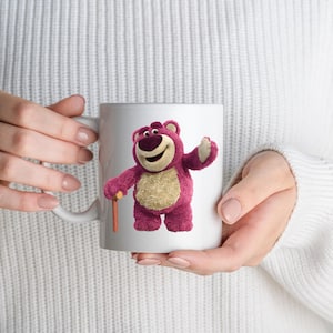 May include: White ceramic mug featuring a cartoon image of a pink bear with a friendly expression. The bear is holding a wooden cane. The mug is held by a person wearing a white sweater. The mug is a standard size.