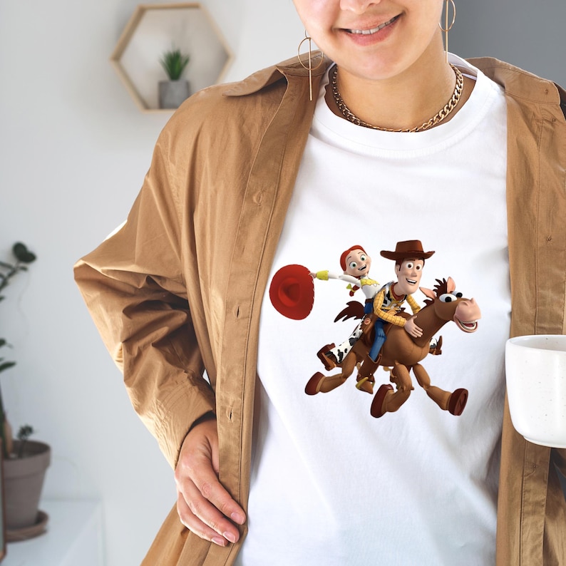 May include: White t-shirt featuring a colorful graphic of Woody and Jessie from Toy Story riding a horse. The design includes a red lasso and a brown cowboy hat. The shirt is worn by a person.