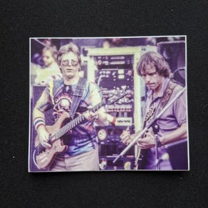 May include: A rectangular sticker featuring a photograph of two musicians performing on stage. The musician on the left wears a tie-dye shirt and glasses while playing an electric guitar. The musician on the right plays a guitar in a purple shirt.