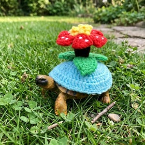 Puede incluir: Una tortuga con una cubierta de caparazón de ganchillo azul claro y una flor de ganchillo en la espalda. La flor tiene pétalos rojos con puntos blancos, detalles amarillos y hojas verdes. La tortuga está sobre hierba verde.