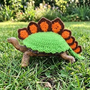 Puede incluir: Una figura de tortuga con un caparazón verde tejido y una espalda de estegosaurio tejida marrón y naranja. La tortuga está en un entorno exterior cubierto de hierba.