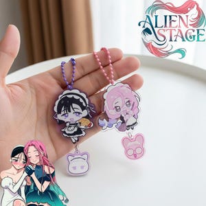 Op de afbeelding: Twee sleutelhangers in anime-stijl met stripfiguren in dienstmeisjesoutfits. De ene sleutelhanger heeft een paarse ketting en een zwart-wit karakter, de andere heeft een roze ketting en een roze karakter. Elke sleutelhanger heeft een bedel met een katachtig gezicht.