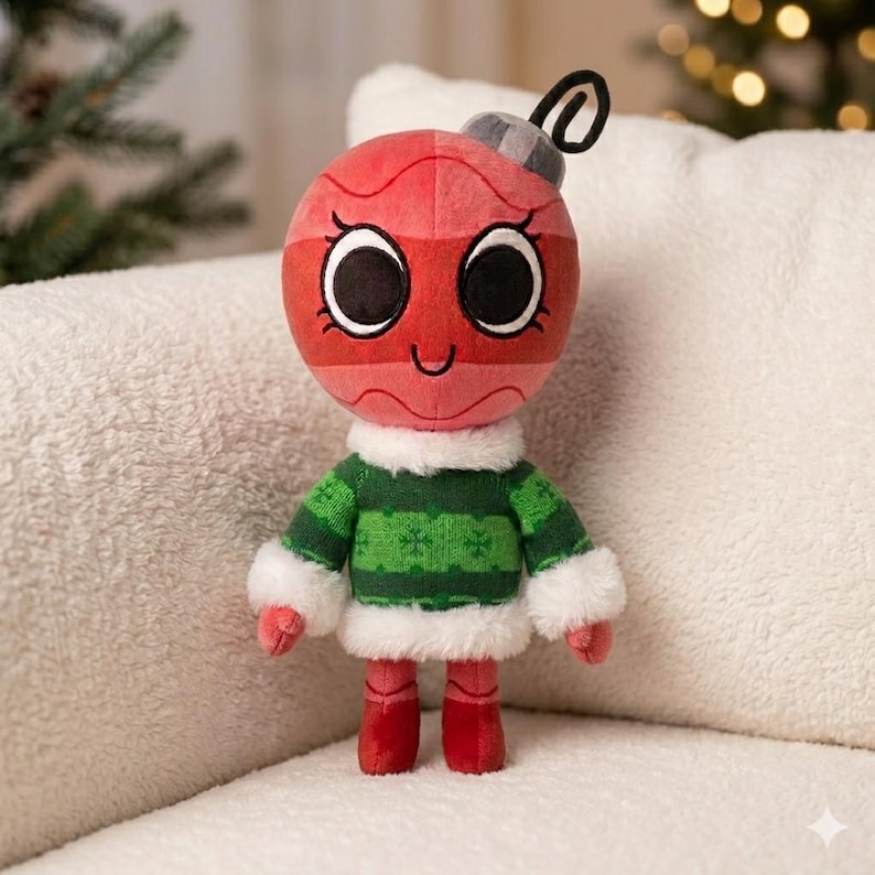 May include: A festive plush Christmas ornament character. It features a red body, large black eyes, and a smiling expression. The character is dressed in a green sweater with white trim and red boots, and has a silver cap.