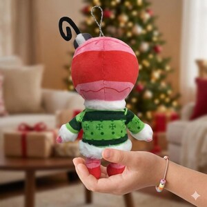 May include: A plush Christmas ornament shaped like a character, held in a hand. The ornament has a red and pink head, a green sweater with snowflake designs, and red feet. A silver wire loop is attached for hanging.