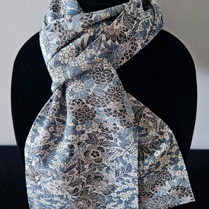 May include: A long scarf featuring a dense, intricate floral pattern in shades of blue, grey, and white. The fabric displays a textured surface with overlapping botanical motifs, including various flower shapes and leaves, creating a continuous, detailed design throughout the entire length of the accessory.
