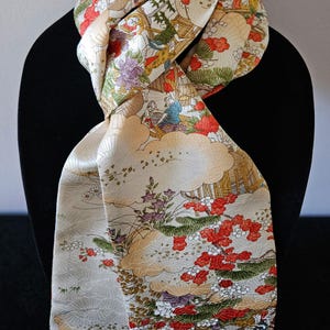 May include: A cream-colored scarf featuring a detailed print of traditional Japanese scenes, including stylized red flowers, green pine branches, and figures in architecture, with gold-toned cloud-like patterns throughout the fabric.