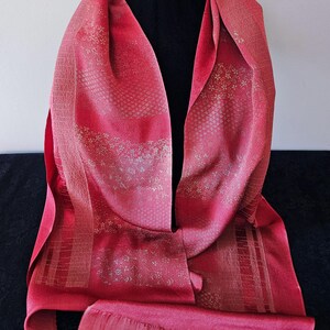 May include: A vibrant red scarf featuring a textured surface with subtle, repeating floral patterns and geometric bands. The fabric displays a slight sheen, with lighter red and gold-toned accents woven into the design, finished with a straight hem.