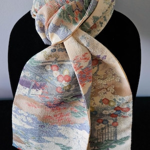 May include: A cream-colored scarf with a detailed floral and landscape pattern in shades of red, blue, green, and purple. The scarf is draped over a black mannequin, showcasing its design and texture. The scarf's fabric appears to have a subtle, textured surface.