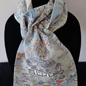May include: A light blue scarf featuring a detailed woven pattern of traditional buildings, trees, and floral elements in shades of white, gray, gold, and red. The fabric has a textured surface and is draped to show the intricate landscape design.