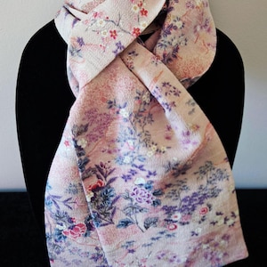May include: A light pink scarf with a floral pattern. The scarf is draped over a black form, showcasing its design. The pattern includes flowers in shades of red, purple, and white, set against a light pink background.