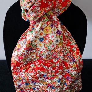 May include: A vibrant red scarf featuring a dense, intricate pattern of multi-colored flowers and green foliage. The fabric has a smooth, slightly reflective finish, and the scarf is tied in a simple knot, displaying a rich array of white, yellow, blue, and purple floral details across the entire surface.