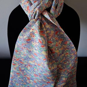 May include: A light purple scarf with a repeating pattern of colorful fans and floral designs. The scarf is draped over a black form, showcasing its intricate details and vibrant colors. The scarf's fabric appears soft and lightweight.