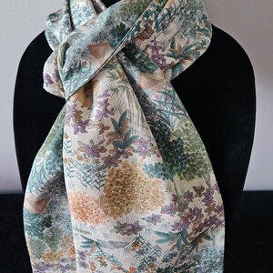 May include: A silk scarf with a botanical print in shades of green, purple, and beige. The scarf is draped over a black mannequin head, showcasing its intricate floral and foliage design. The scarf's pattern includes trees, flowers, and other natural elements.