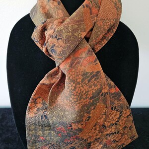May include: A long scarf featuring a textured fabric with a dense, multi-colored pattern of orange, brown, and dark green floral and geometric motifs. The scarf is tied in a simple knot, displaying a complex, woven-style design throughout.