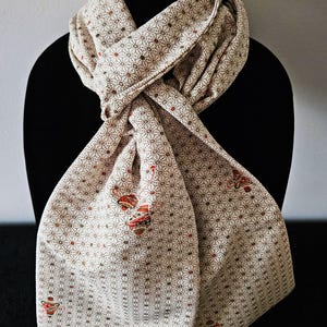 May include: Cream-colored scarf with a repeating geometric pattern and red and brown accents. The scarf is draped over a black form, showcasing its intricate design and soft texture. The scarf features a detailed floral pattern.