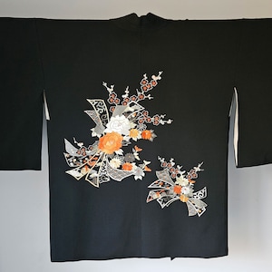 May include: Black kimono with floral embroidery. The design features white, orange, and gray flowers with decorative ribbon elements. The kimono is displayed on a black metal rack.
